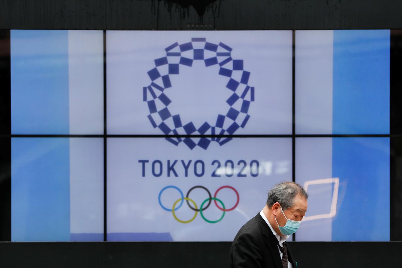 A passerby wearing a protective face mask walks past a screen showing the logo of the Tokyo 2020 Olympic Games in Tokyo