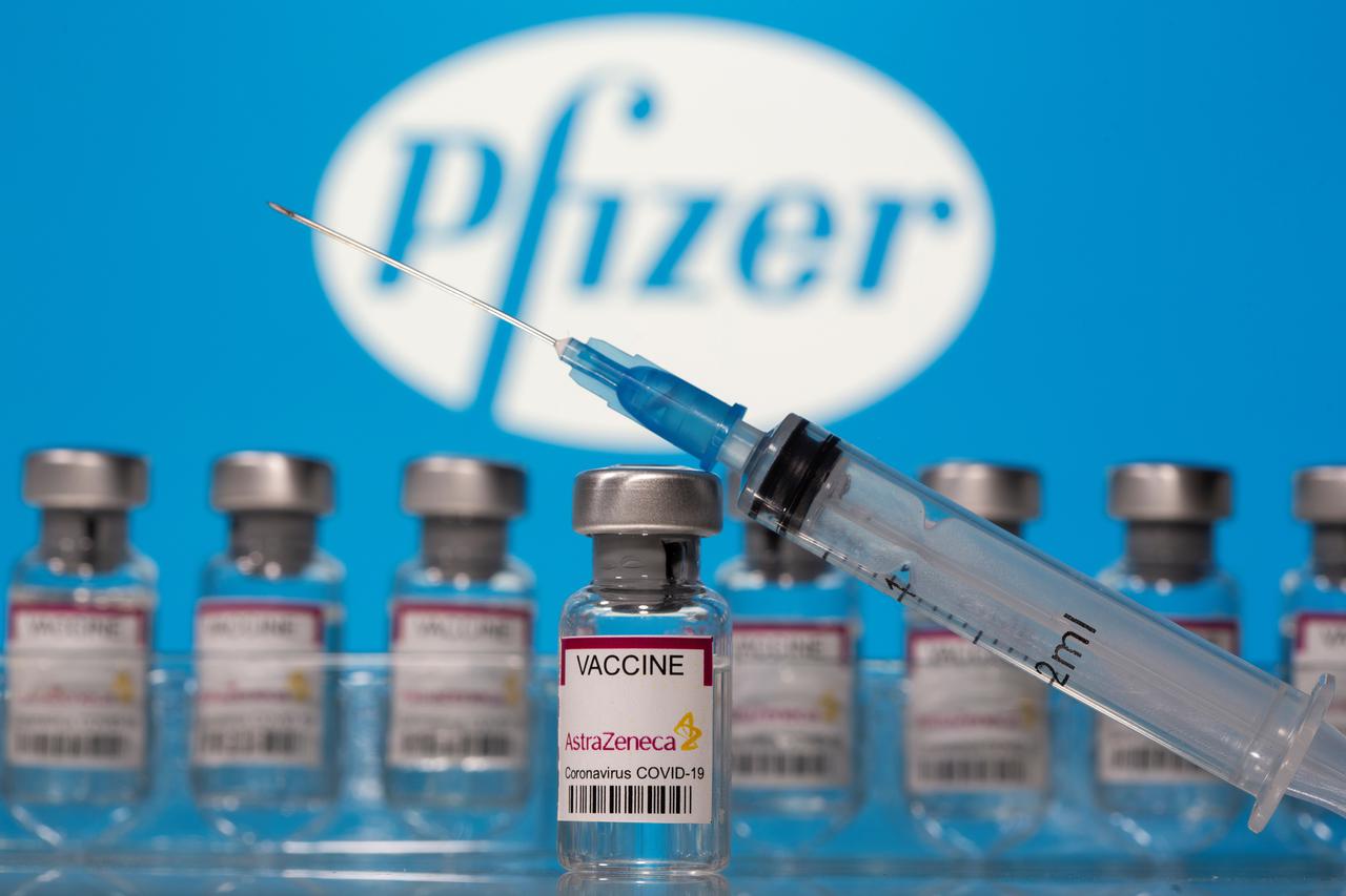 Vials labelled "AstraZeneca COVID-19 Coronavirus Vaccine" and a syringe are seen in front of a displayed Pfizer logo