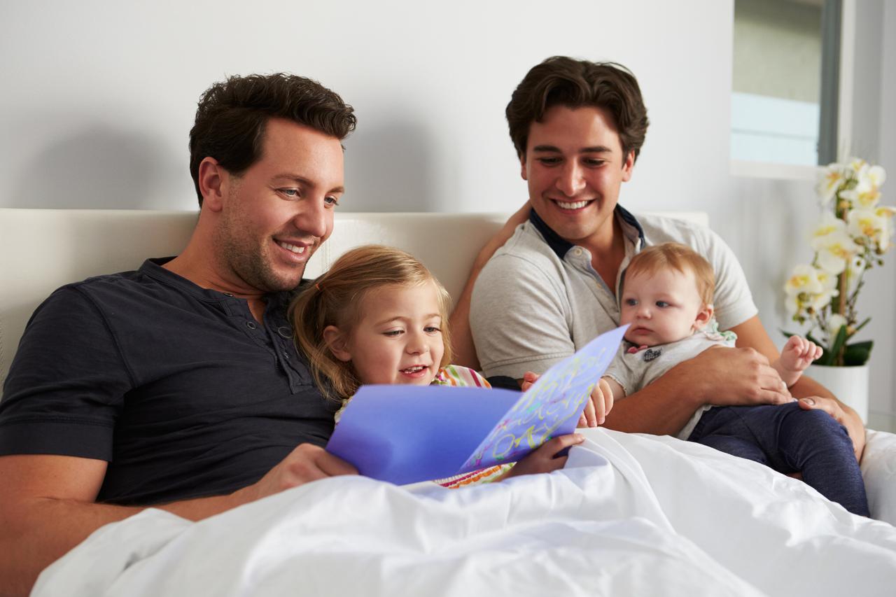 Male gay parents relaxing in bed with two kids