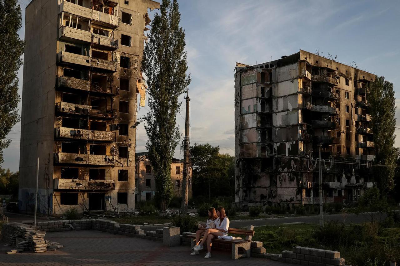 Two girls sit in a public square in front of destroyed buildings in Borodianka