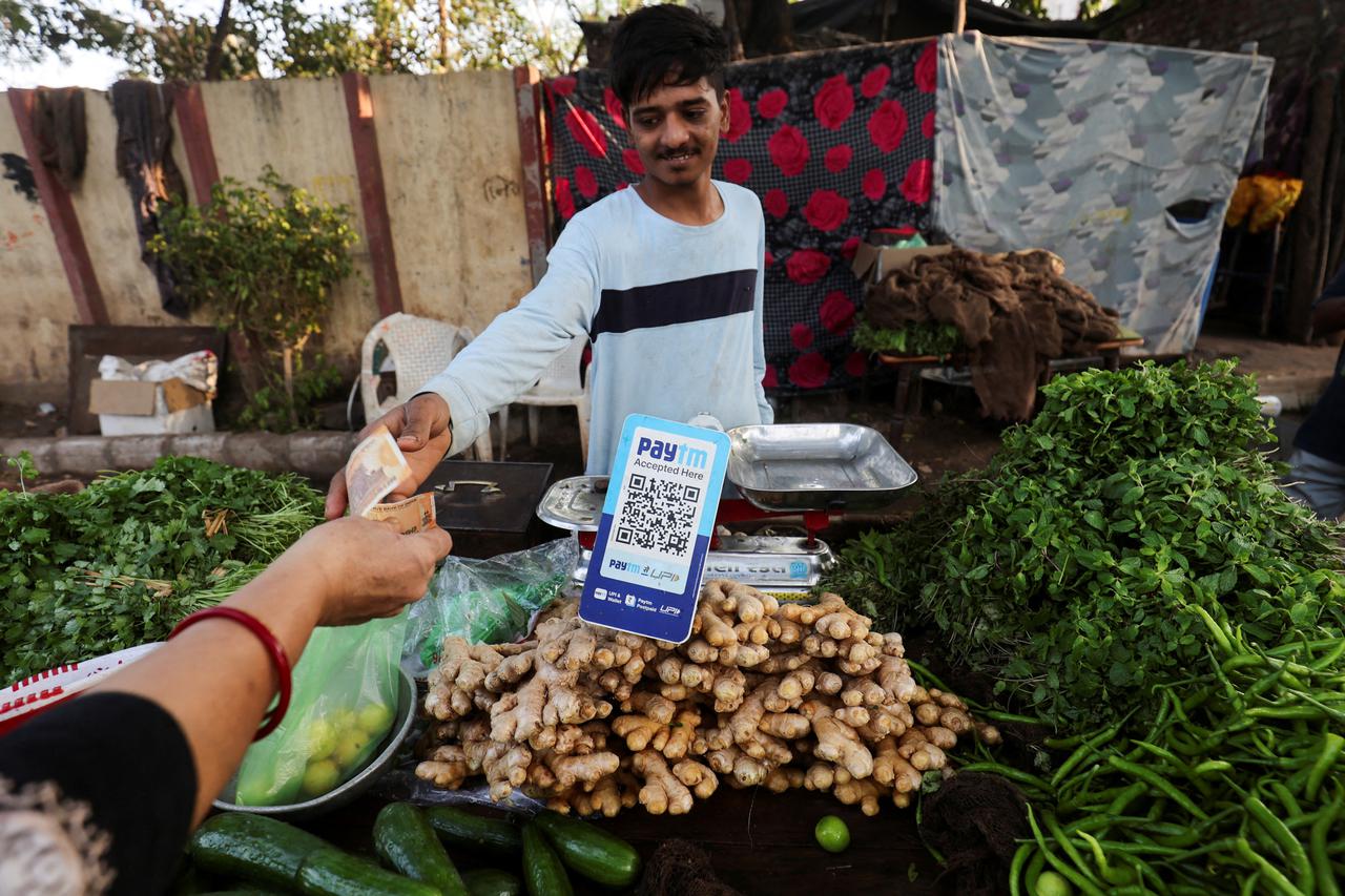 FILE PHOTO: A customer pays cash to buy vegetables next to a QR code of Paytm, a digital payments firm, on display at a roadside market in Ahmedabad