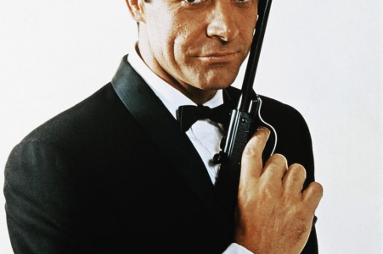 'ca. 1968 --- Waist-up portrait of Sean Connery, as James Bond, caressing the barrel of a gun against the side of his face.  Connery is wearing a tuxedo and bow tie and smiling slightly. --- Image by 